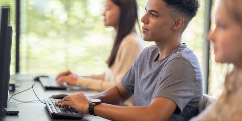 A imagem mostra três adolescentes sentados em frente a computadores digitando. Em primeiro plano, está um menino pardo que veste camiseta cinza e usa relógio no punho. Em segundo plano, desfocadas, estão uma menina loira com blusa branca de manga comprida e uma menina oriental de blusa de lã branca.