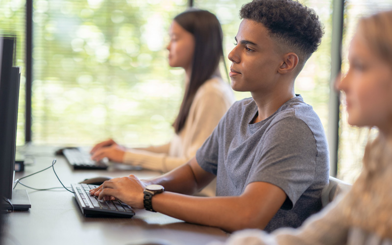 A imagem mostra três adolescentes sentados em frente a computadores digitando. Em primeiro plano, está um menino pardo que veste camiseta cinza e usa relógio no punho. Em segundo plano, desfocadas, estão uma menina loira com blusa branca de manga comprida e uma menina oriental de blusa de lã branca.