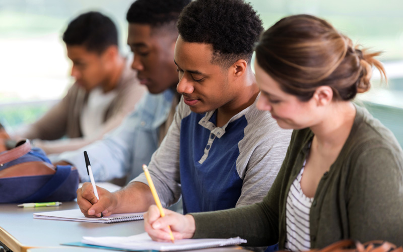 Imagem mostra grupo de estudantes em sala de aula, sentados, com canetas e lápis em mãos, escrevendo em cadernos. Vemos estudantes homens negros e uma estudante mulher branca