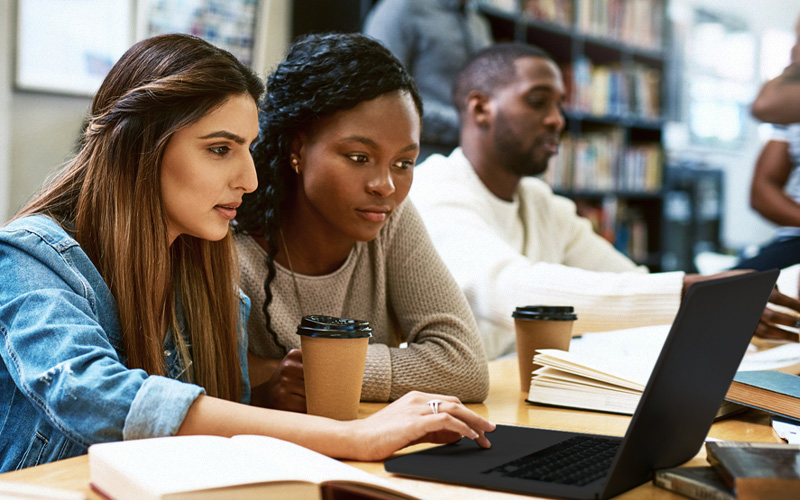 Imagem mostra três jovens em uma sala de estudos utilizando notebook. Dois jovens são negros e uma é branca