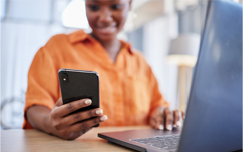 Imagem mostra uma professora negra segurando um celular com a mão direita. Há um notebook sobre a mesa
