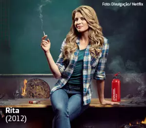 Professora Rita aparece sentada em uma mesa e fumando cigarro em cena da série da Netfilx