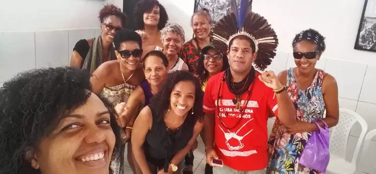 Integrantes do coletivo feminista da Feira das Mulheres de Itapuã estão sorrindo para a câmera. Em primeiro plano, uma mulher de cabelos encaracolados está tirando a selfie e um dos integrantes usa cocar.