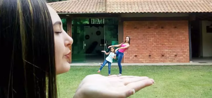 Imagem mostra menina “gigante” “assoprando” duas outras meninas na mão (ilusão de perspectiva). Ao fundo se vê uma casa.