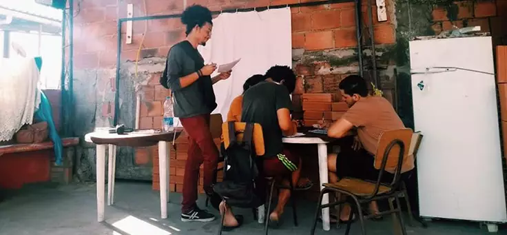 Jovem com cabelos crespos presos em rabo de cavalo orienta outras três jovens sentados em carteiras no projeto Unifavela, que contribui para a formação de jovens.