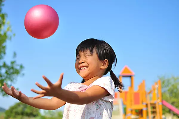 A imagem mostra uma criança jogando uma pequena bola rosa para cima. Ela sorri com os olhos fechados.
