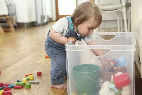A imagem mostra uma criança pequena colocando a mão dentro de uma caixa de plástico transparente onde estão vários objetos