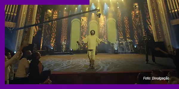 Imagem mostra cena do espetáculo AmarElo. O rapper Emicida está no palco do Theatro Municipal de São Paulo. Ele usa roupa na cor amarela e está de braços abertos. Está acompanhado de músicos no palco. Parte do público aparece na imagem.