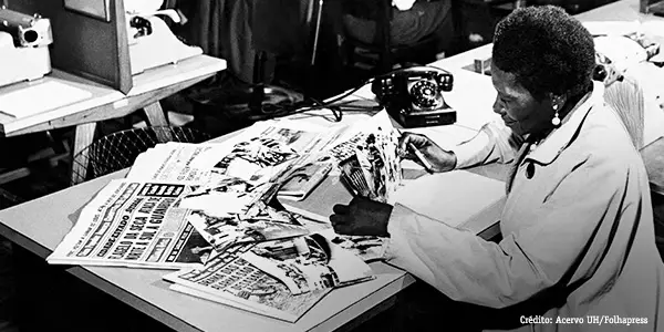 Imagem mostra a escritora lendo diversos jornais que estão espalhados em cima de uma mesa.